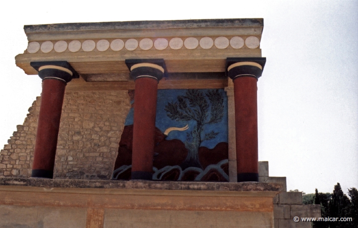 9533.jpg - 9533: Bull Bastion on the west side of the Palace of Knossos. An open air passage linked the Central Court with the North Entrance. It was paved and sharply inclined towards the north. The passage is narrow. Right and left were two raised colonnades known as “Bastions.” Arthur Evans reconstructed the “Bastion” on the west side. He also placed a copy of a restored relief fresco of a bull here. The wall painting may have formed part of a hunting scene. The passage ends in a large hall with ten square pillars and two columns. The pillars and columns probably supported a large hall on the upper floor. Evans suggested that, due to its position on the seaward side, it was here that the produce of seaborne trade would have been checked when it reached the Palace. It was therefore named the “Customs House”. Palace of Knossos (Crete).